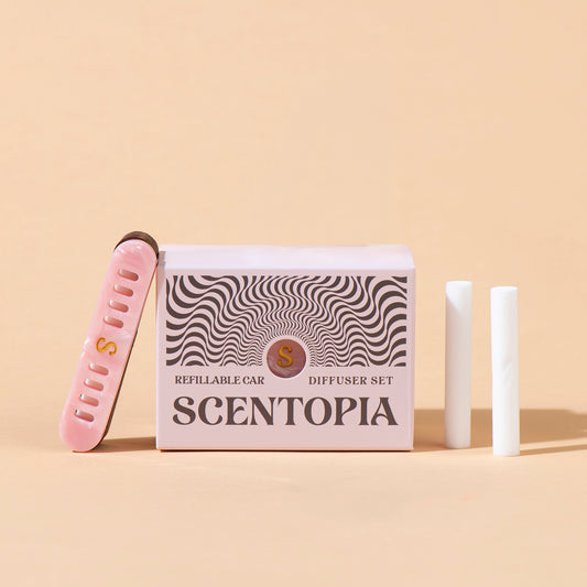 Scentopia Petal refillable car diffuser set featuring a pink box with black wavy patterns, a pink nail file, and two white car diffuser rods on a beige background.
