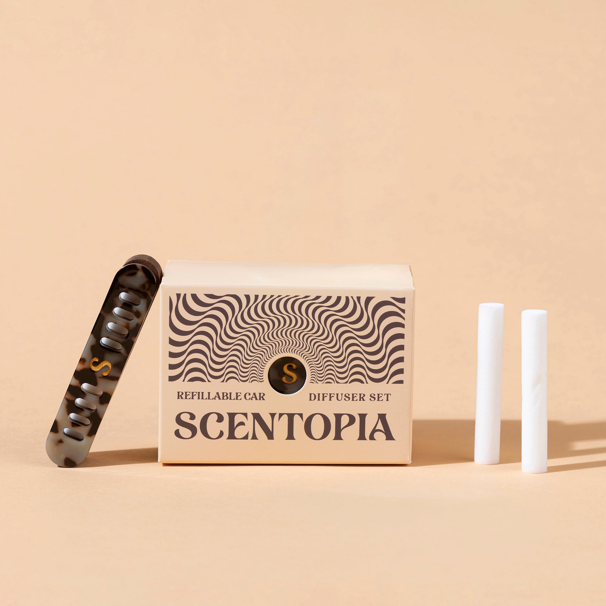 Scentopia refillable car diffuser set featuring a beige patterned box, a tortoiseshell aceate car diffuser, and two white refil rods on a beige background.