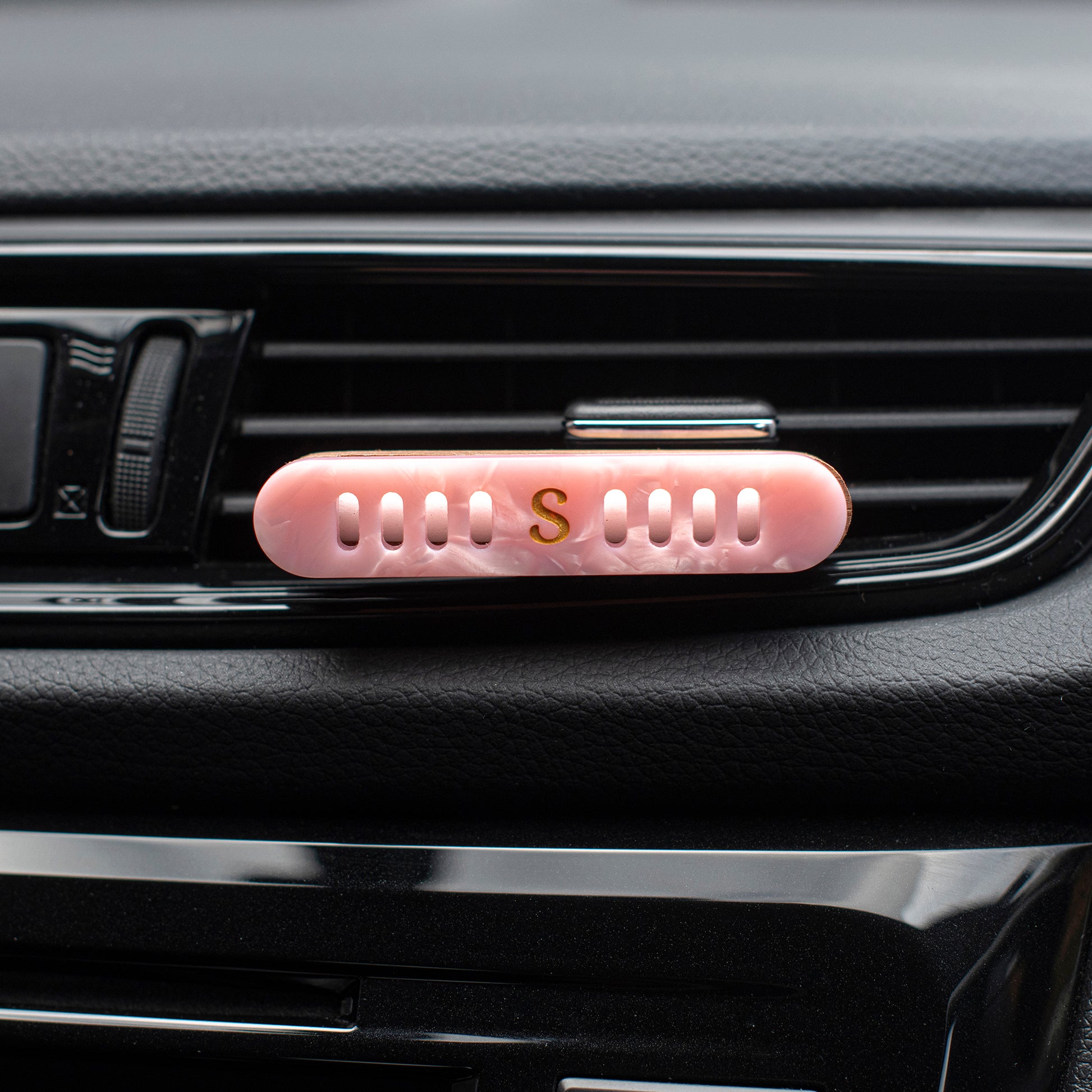 Scentopia petal pink car air freshener diffuser with a gold letter 'S' on the front, clipped onto a black car air vent.