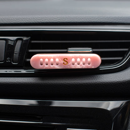 Scentopia petal pink car air freshener diffuser with a gold letter 'S' on the front, clipped onto a black car air vent.