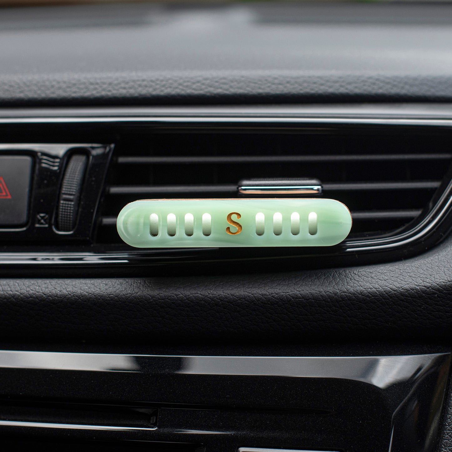 A  Scentopia light jade green car vent diffuser with a small gold letter 'S' in the center, clipped onto a black car air vent.