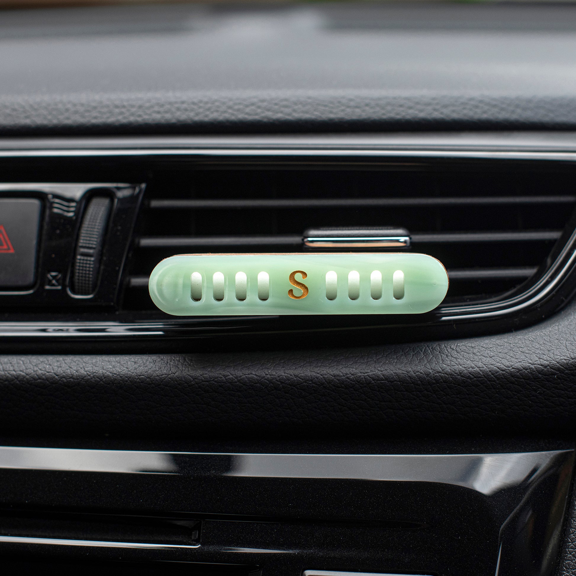 A  Scentopia light jade green car vent diffuser with a small gold letter 'S' in the center, clipped onto a black car air vent.