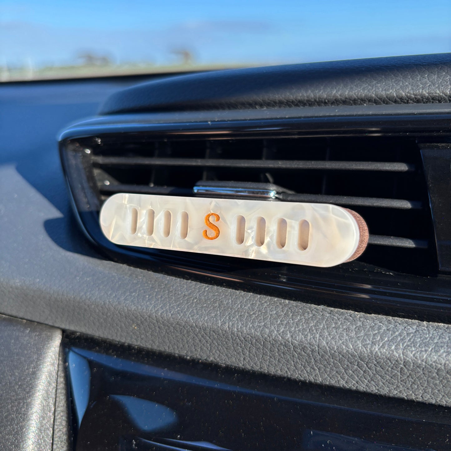 A close-up of a Scentopia car diffuser with a pearl white oval decorative lid featuring a prominent gold letter 'S' in the center, mounted on the car vent .