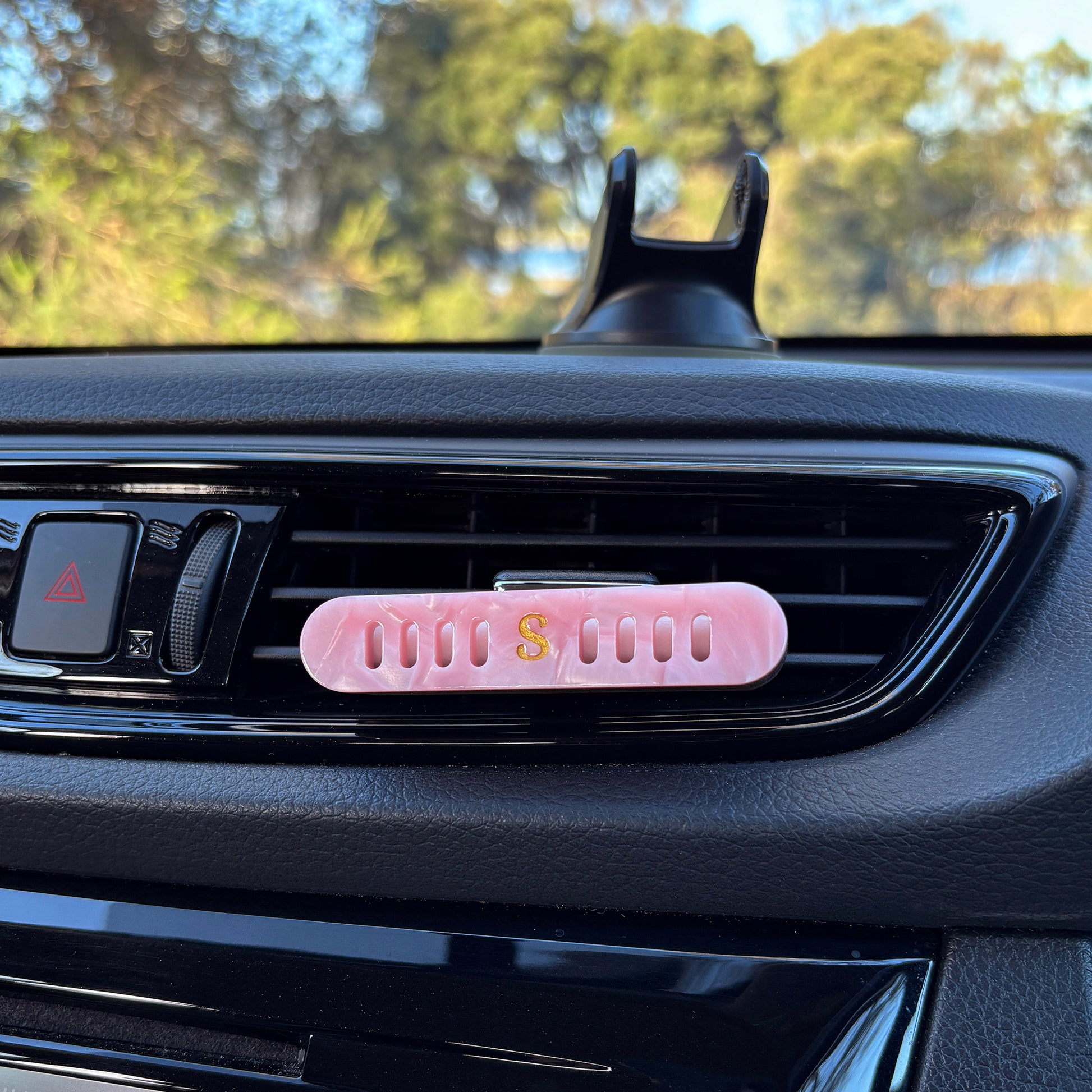Scentopia petal car air freshener diffuser with a small gold 'S' logo, placed on a black car air vent with trees visible in the background.