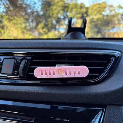Scentopia petal car air freshener diffuser with a small gold 'S' logo, placed on a black car air vent with trees visible in the background.