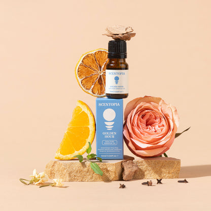 A Scentopia Golden Rose essential oil bottle and matching blue box are styled on a stone base with orange slices, a peach rose, and small flowers against a warm beige background.