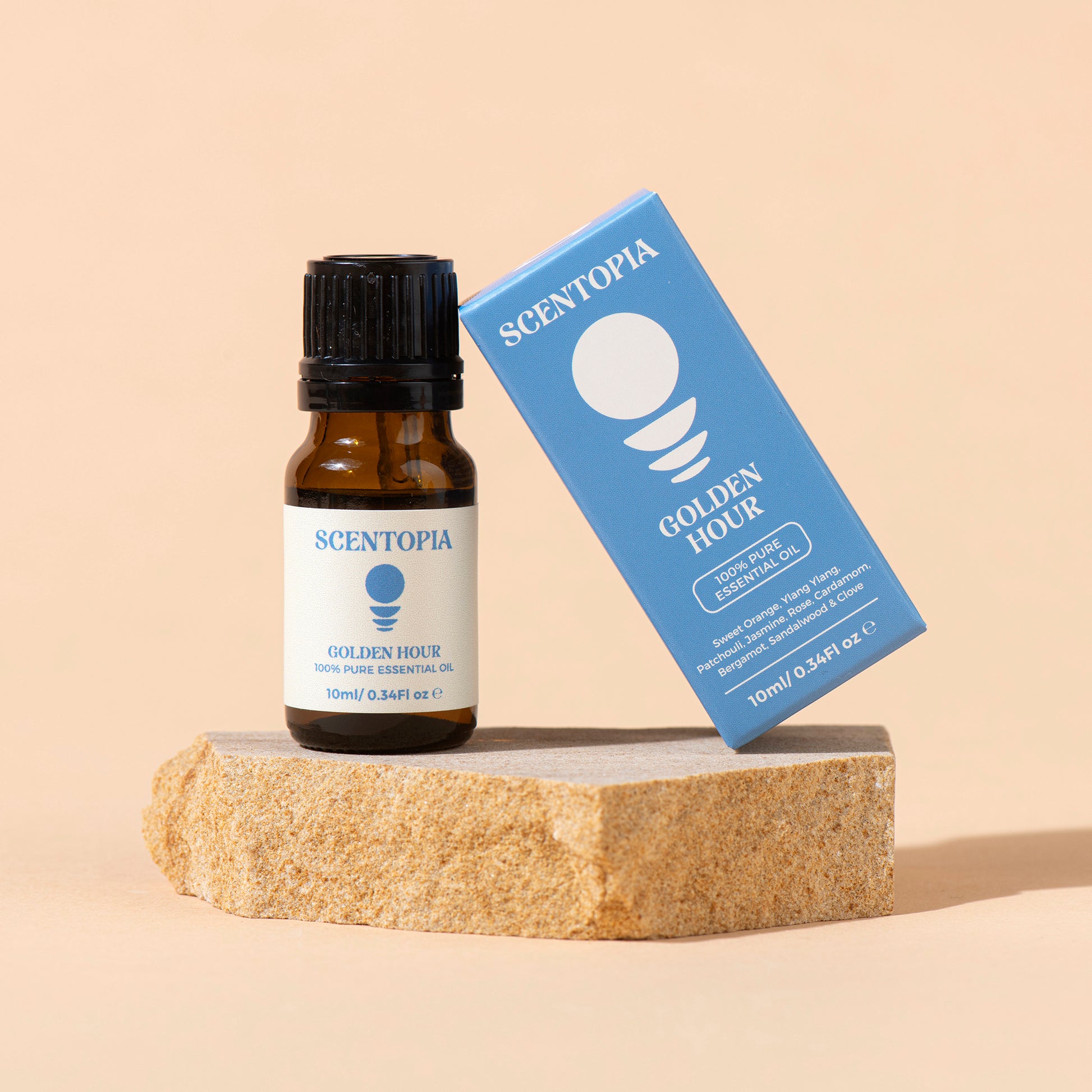 Scentopia Golden Hour 10ml pure essential oil bottle with a black cap next to its blue packaging box, displayed on a textured stone block against a warm beige background.