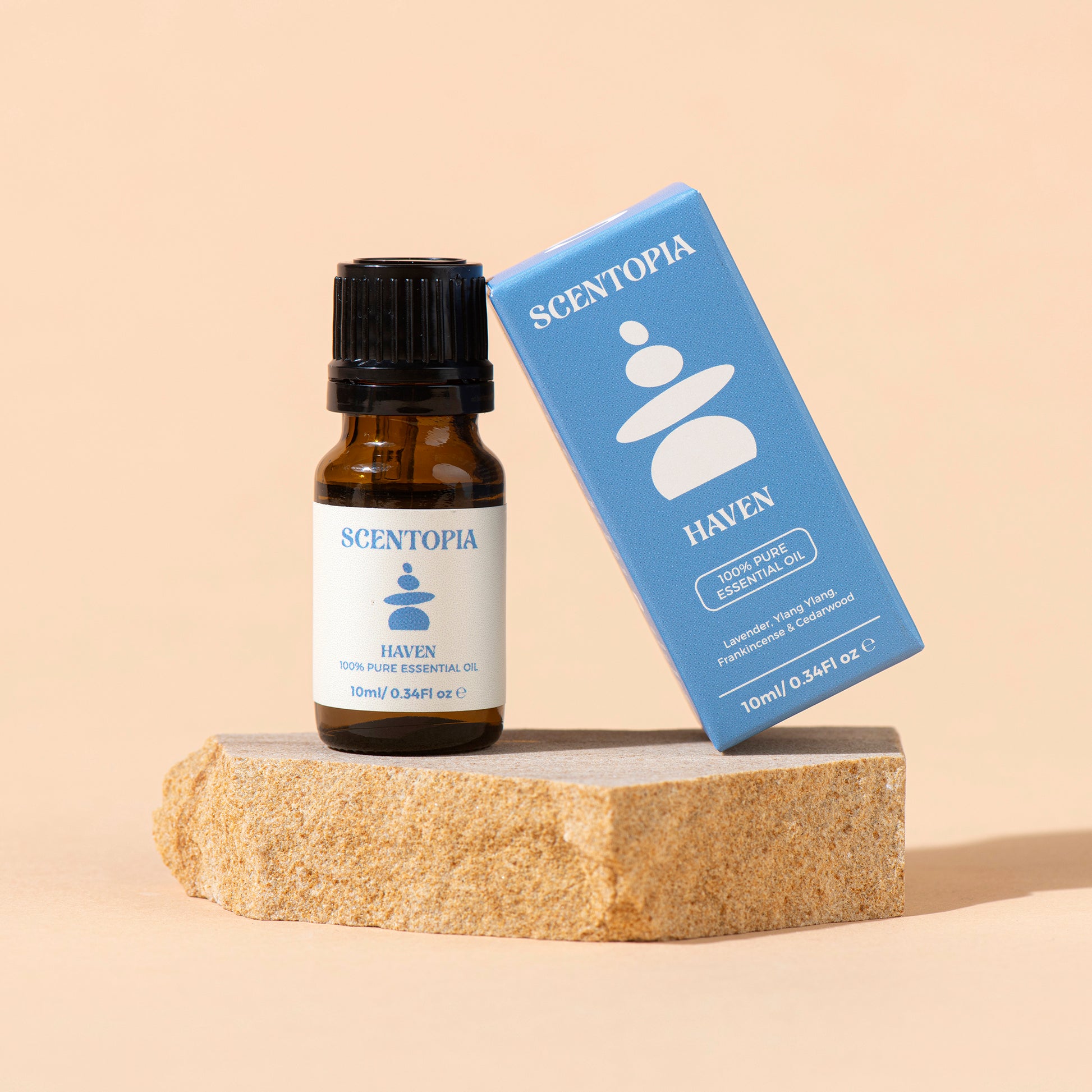 A small amber glass bottle of Scentopia Haven 100% pure essential oil with a black cap stands next to its blue packaging box on a textured stone slab against a warm beige background.