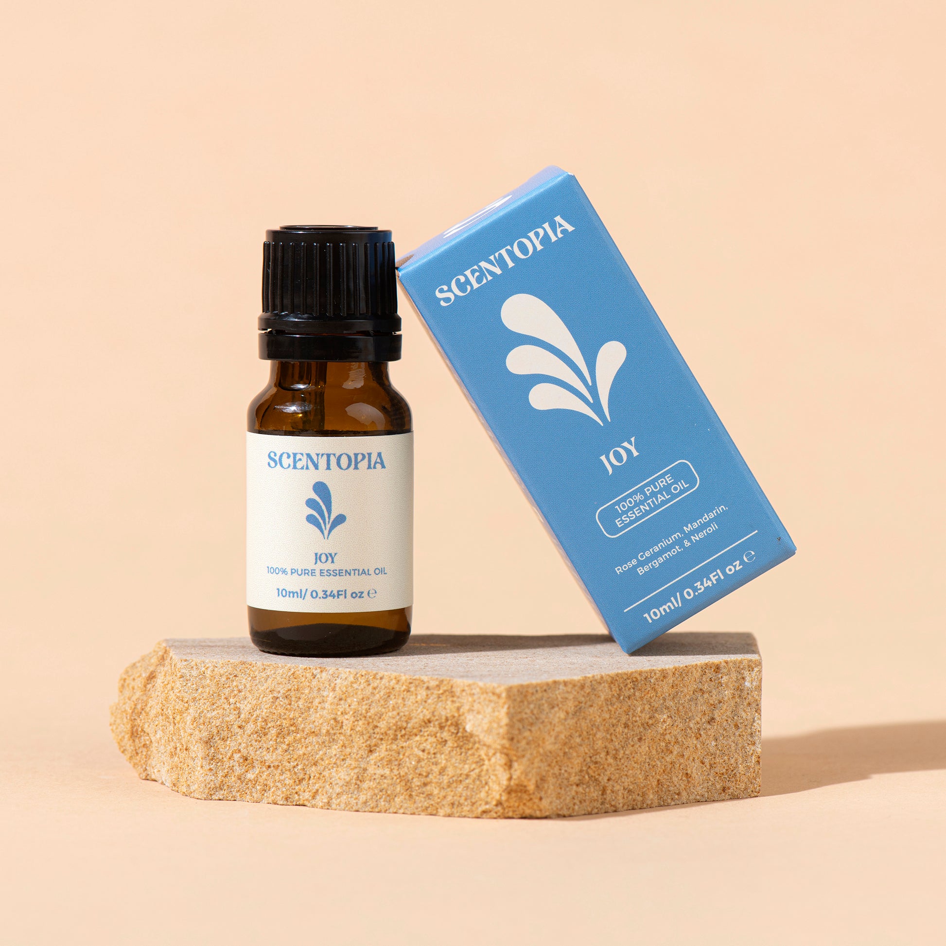A bottle of Scentopia Joy 100% Pure Essential Oil next to its blue packaging box, displayed on a textured stone block against a solid beige background.