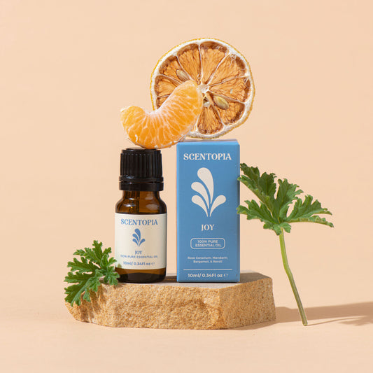 Scentopia Joy 100% Pure Essential Oil bottle and box displayed on a stone slab with dried orange slices, a peeled orange segment, and green leaves against a warm beige background.