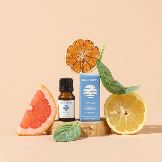 A small amber bottle and matching blue box labeled SCENTOPIA REVIVAL are arranged on a wooden block with citrus slices and basil leaves against a warm beige studio background.