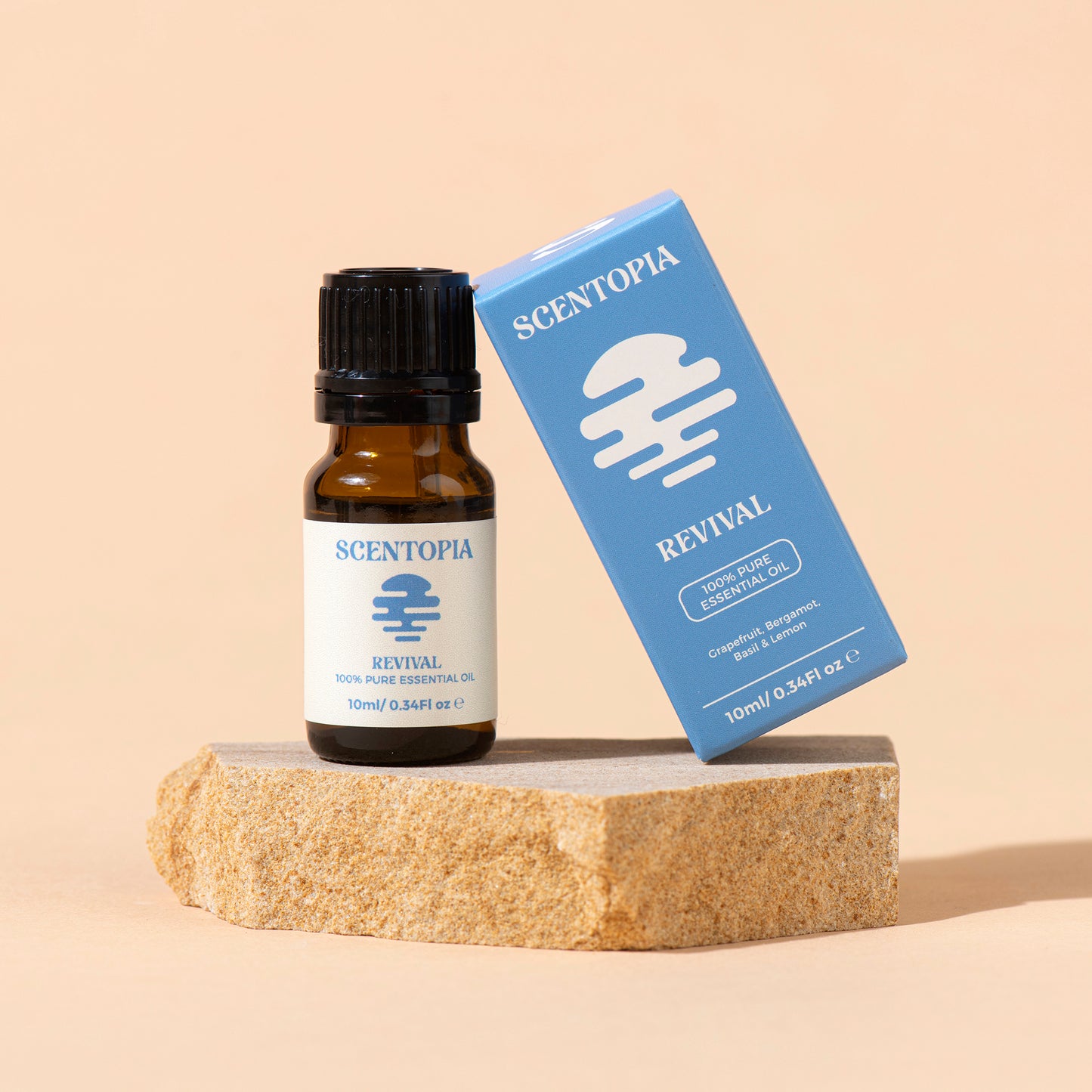 A small amber glass bottle of Scentopia Revival 100% pure essential oil stands next to its blue box on a textured stone slab against a warm beige background.